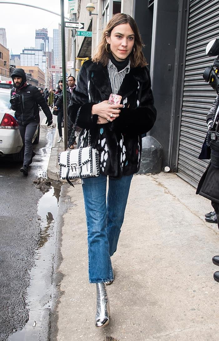 A modelo Alexa Chung, uma mulher branca e jovem, de cabelo ondulado curto, andando pelas ruas de Nova York. Ela usa uma camisa de botão branca, um casaco preto, calça jeans e uma bolsa branca de couro, modelo PS1, da marca Proenza Schouler. - Metrópoles
