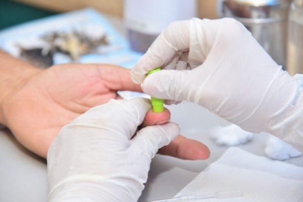 Foto de pessoa com luvas cirúrgicas fura dedo de outra pessoa em exame - Metrópoles