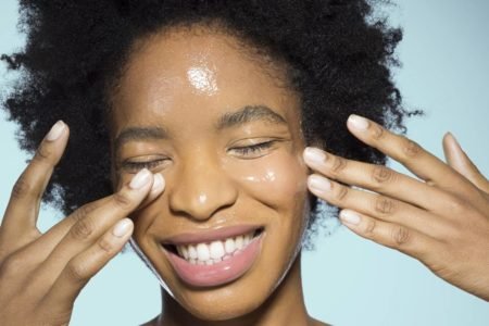 Mulher negra de olhos fechados enquanto passa um produto de textura aquosa no rosto com as duas mãos