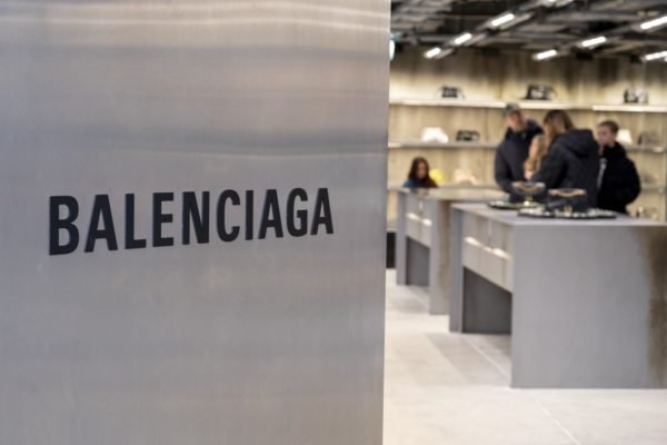 Loja da marca Balenciaga em dezembro de 2022, em Londres, na Inglaterra. Na imagem é possível ver uma porta metálica e a logo da marca em preto. Ao fundo estão prateleiras com produtos e clientes olhando as peças. - Metrópoles
