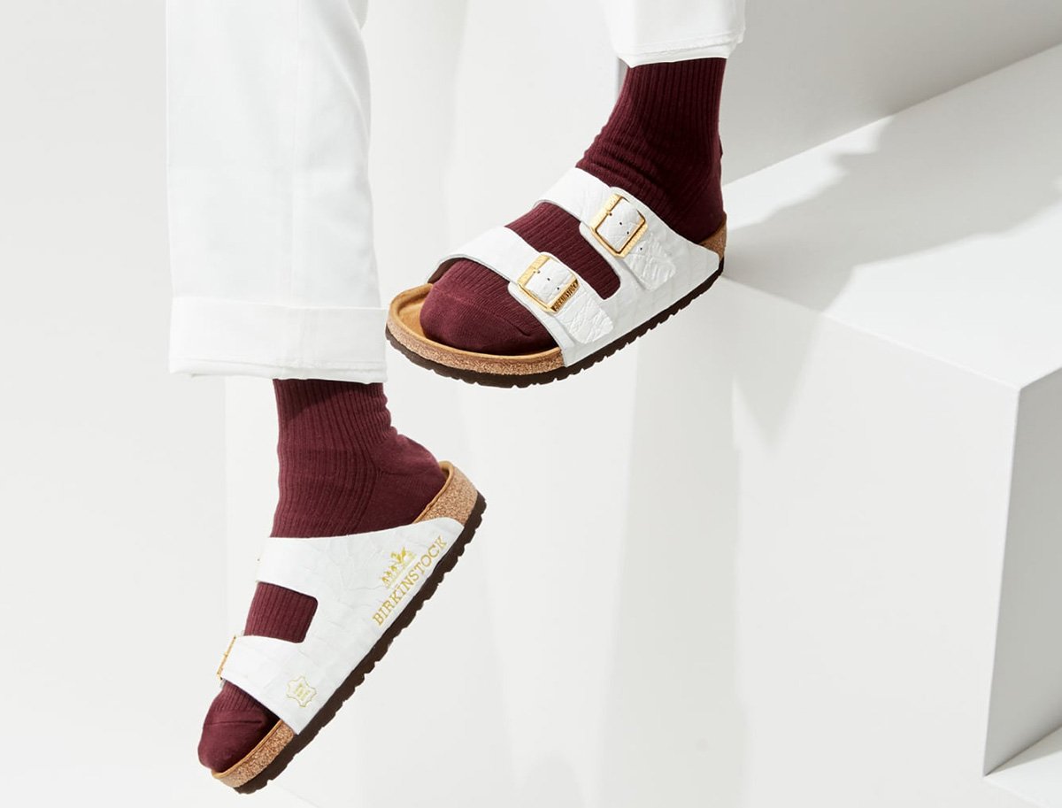 Campanha de divulgação da sandália Birkinstock, da marca MSCHF. O calçado é uma sandália baixa, feita de cortiça e couro. Na foto, a pessoa usa a sandália, na versão de couro branco, com uma meia na cor vinho. - Metrópoles