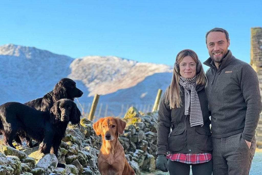Foto colorida de três cachorros ao lado de mulher branca, loira ao lado de homem branco. Eles estão em cima de uma colina - Metrópoles
