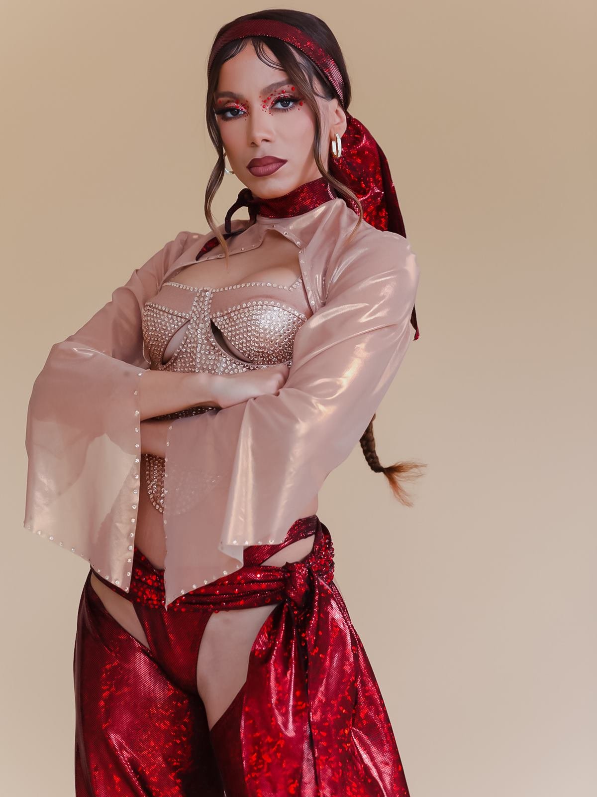 A cantora Anitta, uma mulher parda e jovem, de cabelo ondulado castanho, posa para foto em um fundo bege. Ela usa uma roupa brilhante, rosa e vermelha, feita inspirada nas roupas da revolucionária Anita Garibaldi. - Metrópoles
