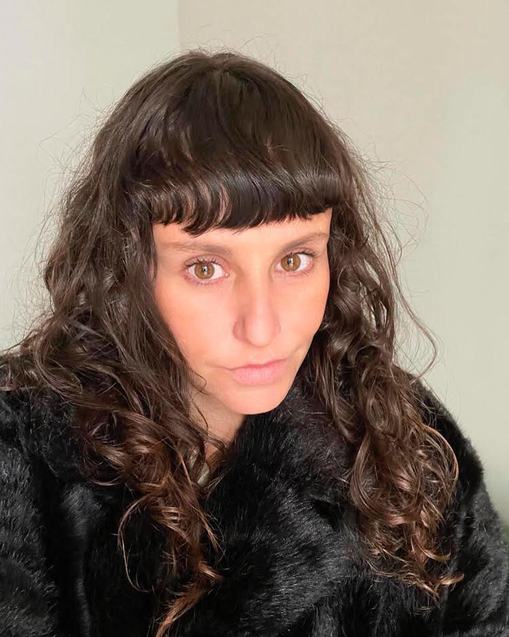 A stylist Clara Lima, uma mulher branca e jovem, de cabelos ondulados castanhos e franja. Ela usa um casaco preto de pelo. - Metrópoles