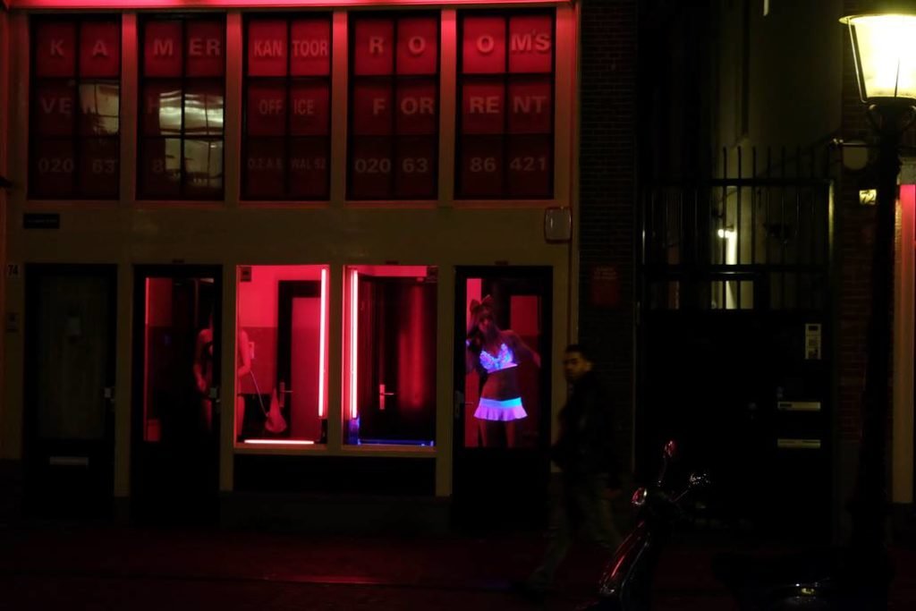 Foto de uma das ruas do Red Light District, em Amsterdã, em que mostra um prostíbulo com luzes vermelhas acesas e uma garota de programa usando lingerie branca que reflete na luz neon