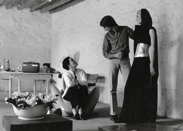 Na imagem em preto e branco, a escultora Claude Lalanne conversa com o estilista Yves Saint Laurent. Ao lado, uma modelo usa look com escultura corporal metalizada - Metrópoles