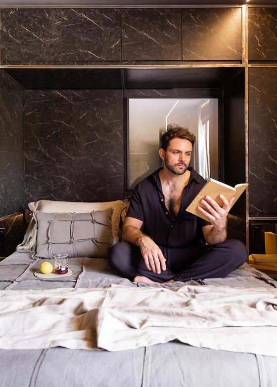 Foto de um homem branco lendo em cima de uma cama de casal