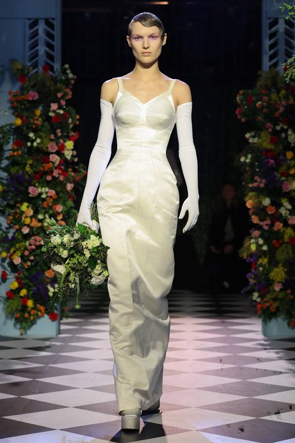 Na passarela, uma modelo usa um vestido de cetim branco com luvas.  Segura um buquê de flores - Metropolis