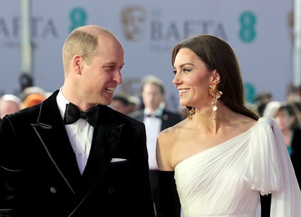 Princesa Kate Middleton usa cabelo partido ao meio e brinco maxi de flores douradas da Zar. Ela usa um longo vestido branco e luvas pretas. Ao lado está o príncipe William com um smoking preto e gravata borboleta. Ele é um homem branco e careca- Zig Proxy 
