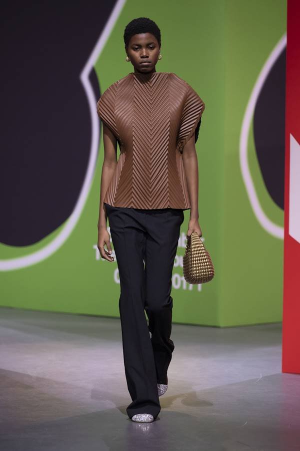 Na passarela, uma modelo usa um look com uma blusa marrom texturizada.  calça de alfaiataria preta e bolsa metálica com esferas - Zig Proxy