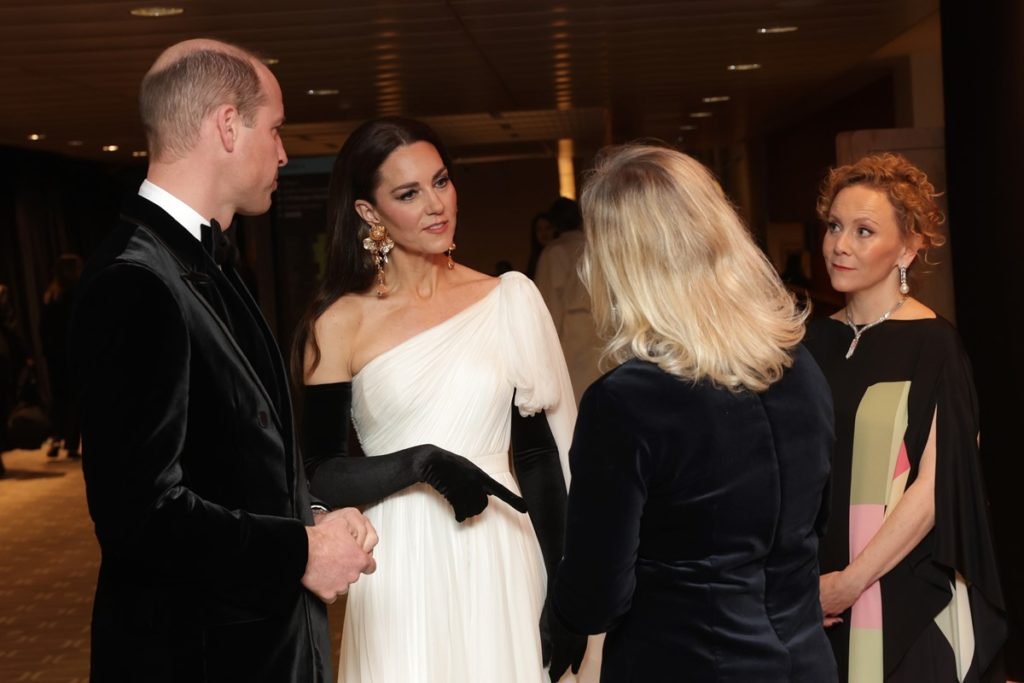Foto colorida de homem branco, calva e com blazer preto ao lado de três mulheres brancas, sendo uma com cabelo preto, outra com fios ruivos e a terceiro com cabelo loiro. Eles estão conversando - Metrópoles