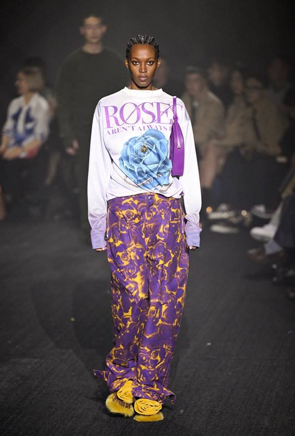 Na passarela a modelo usa look com calça estampada roxa e amarela, além de moletom branco com estampa floral azul - Zig Proxy