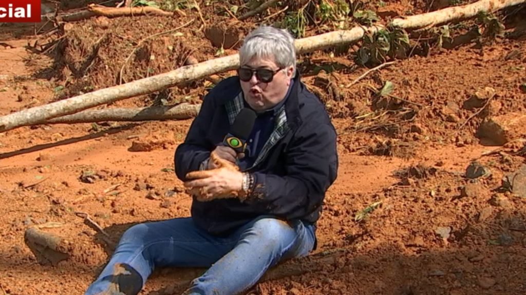 Imagem colorida mostra Datena caido na lama do Litoral Norte de São Paulo. Ele usa óculos escuros e segura o microfone