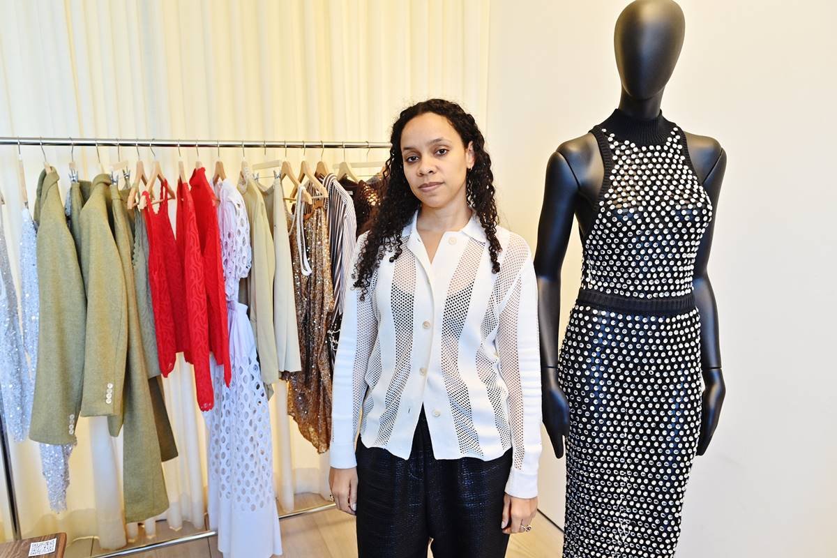 A estilista Rachel Scott, da marca Diotima, posa para foto em um showroom entre uma arara de roupas e uma manequim. Ela é uma mulher negra e jovem, de cabelo preto cacheado, e usa uma camisa de botão branca. - Metrópoles