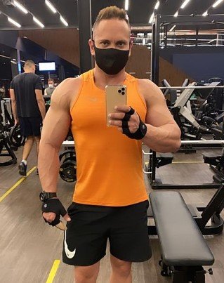 Homem com camisa laranja e máscara preta tirando foto no espelho em academia