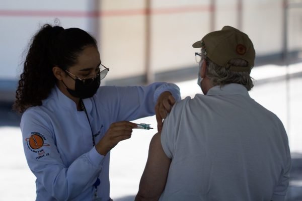 Mulher com máscara e jaleco aplicando vacina em idoso de costas
