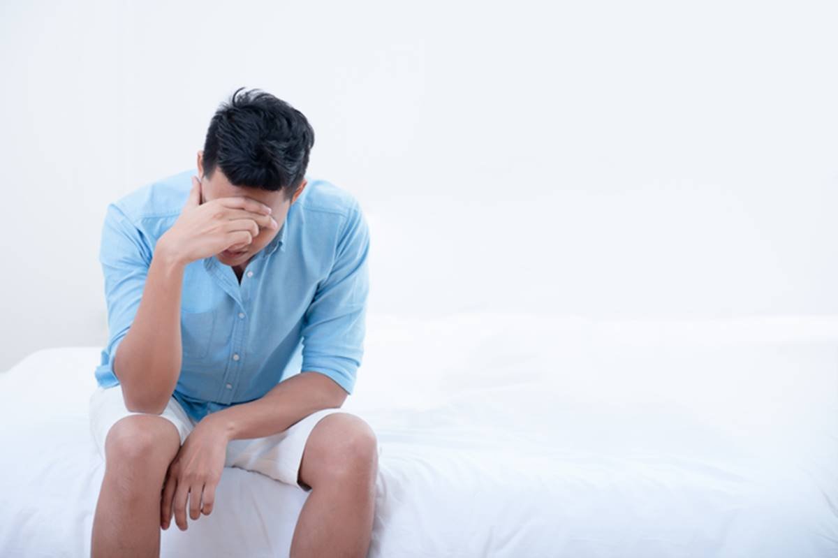 Homem sentado em cama branca com mão escondendo o rosto representando sinal de decepção