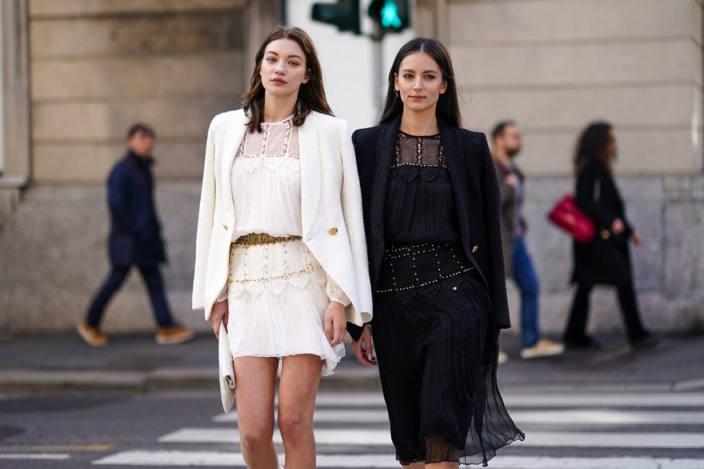Duas amigas mulheres, jovens e brancas, andando pela rua durante a Semana de Moda de Milão. As duas usam roupas iguais, só que em cores diferentes: vestidos folgados, com detalhes de renda, e um blazer por cima. A roupa da primeira é toda branca e, da segunda, toda preta. - Metrópoles