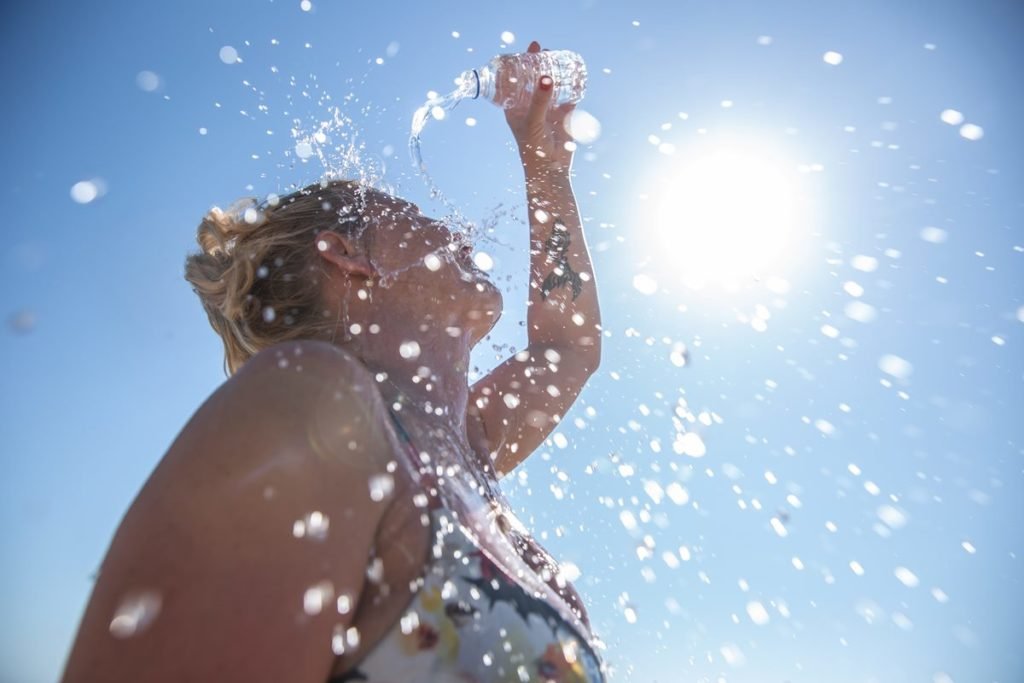 Imagem colorida de mulher ao sol jogando água na cabeça para se refrescar - Metrópoles