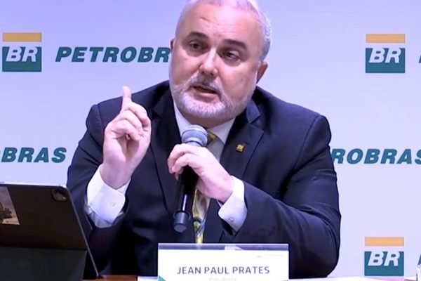 Imagem colorida de Jean Paul Prates, sentado em uma mesa e de terno, em parede com logotipo da Petrobras - Metrópoles