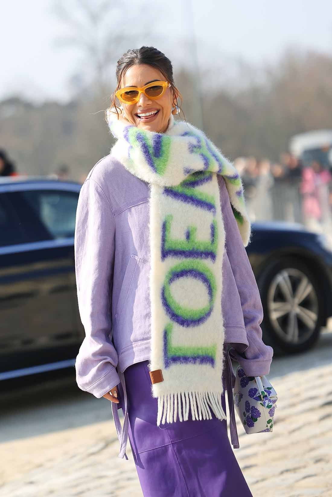 A influencer Camila Coelho no desfile da marca Loewe na Semana de Moda de Paris. Ela é uma mulher branca e jovem, de cabelo liso curto e castanho, e usa uma jaqueta lilás, uma calça roxa e um cachecol branco com o nome da marca Loewe bordado. - Metrópoles