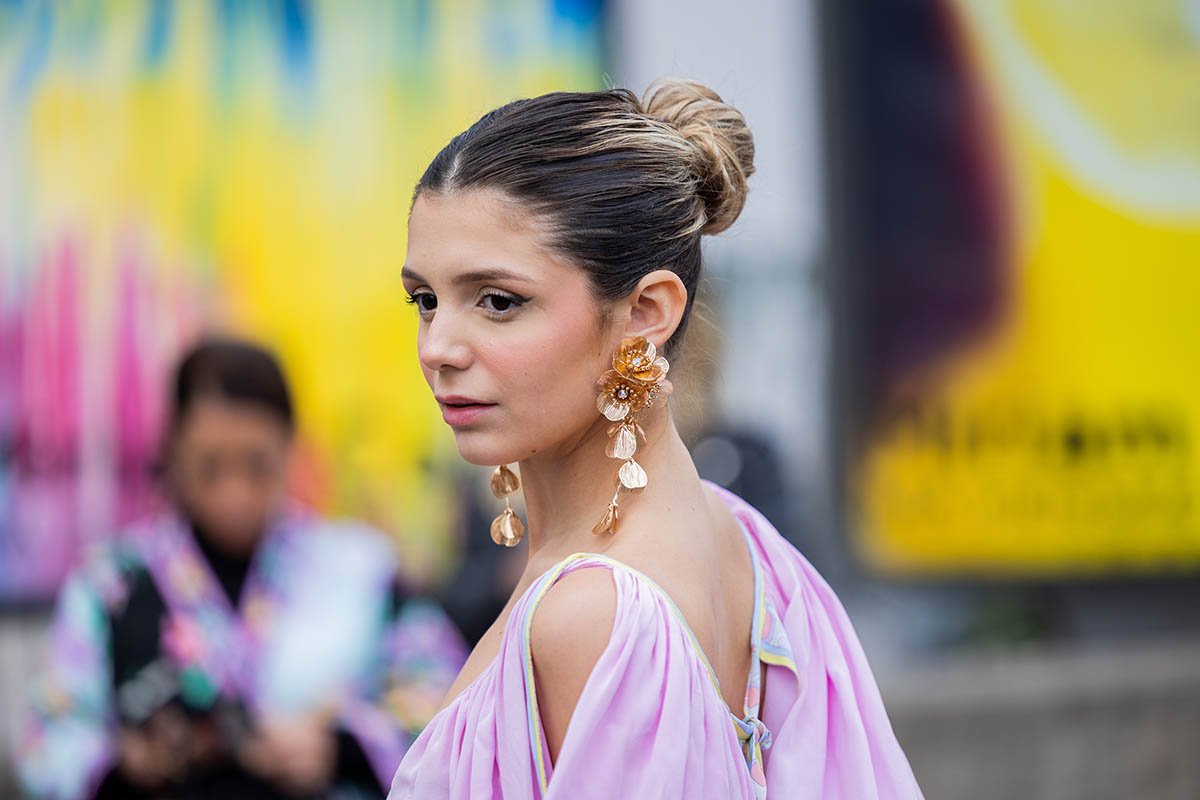 A influencer Catarina Tourinho posa para foto na rua durante a Semana de Moda de Paris. Ela é uma mulher jovem e branca, de cabelo liso loiro, e usa um vestido rosa esvoaçante e brincos grandes em formato de flor na cor dourada. - Metrópoles