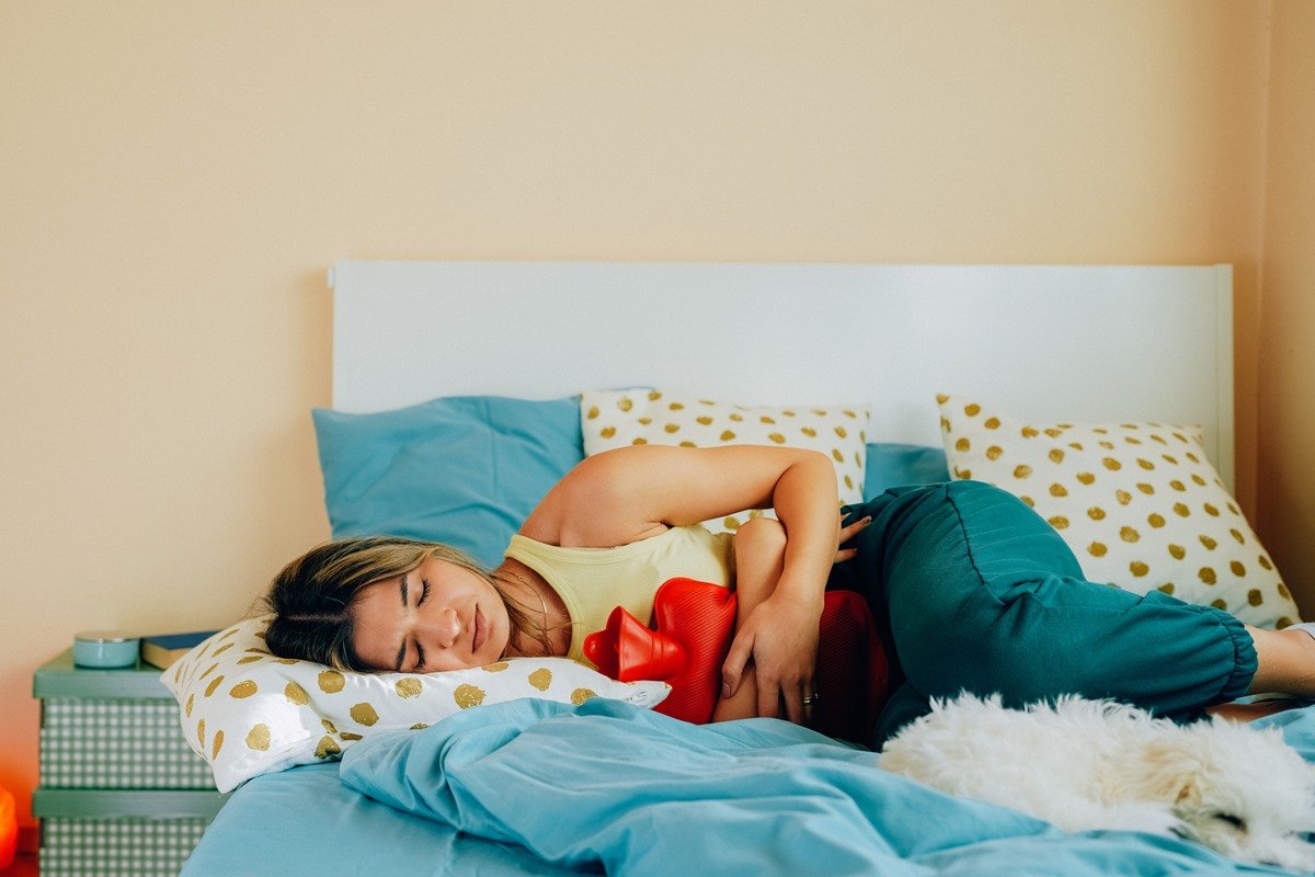 Foto colorida de mulher deitada em cama e com uma bolsa de água quente na barriga - Metrópoles