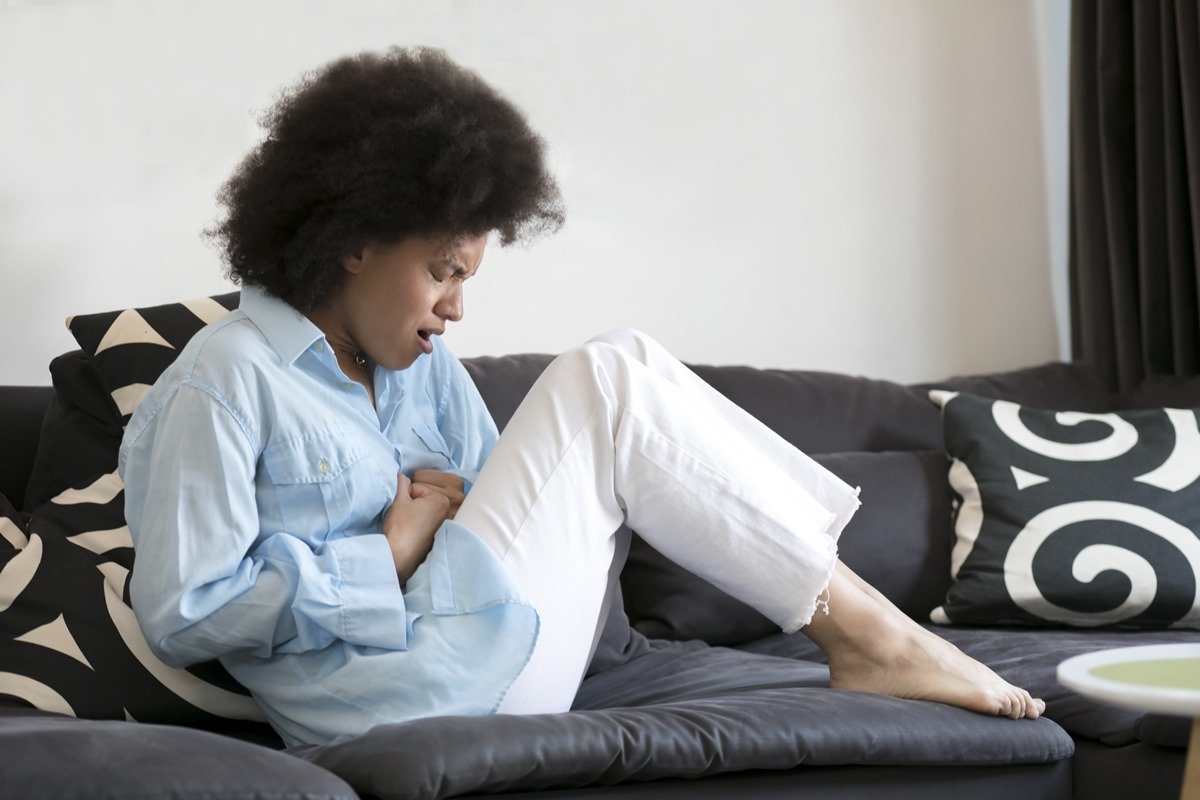 Foto colorida de mulher negra sentada em um sofá sentindo dores na barriga - Metrópoles