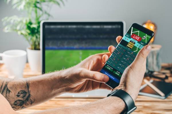 Fotografia colorida mostrando mãos de homem mexendo em celular com televisão ao fundo transmitindo jogo de futebol-Metrópoles