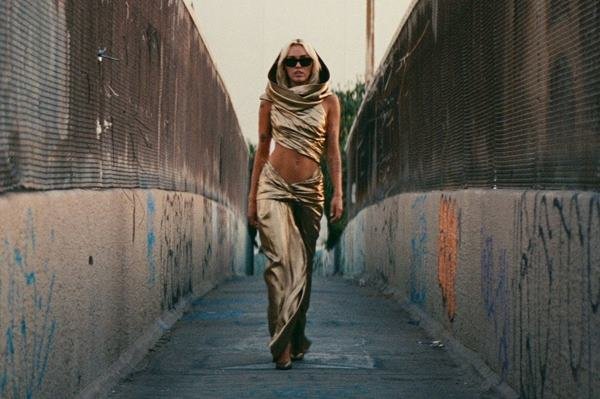 Mulher loira andando em corredor na rua com óculos de sol. Ela está usando vestido dourado com capuz - Metrópoles