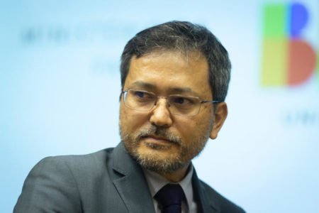 homem de traços orientais, óculos e terno olha para o lado esquerdo
