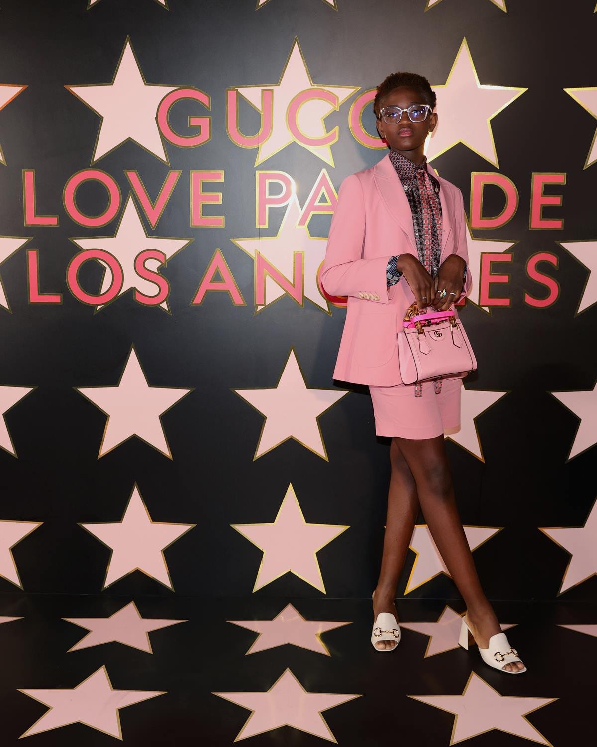 A adolescente Zaya Wade, uma mulher trans e negra, de cabelos trançados pretos, em evendo da marca Gucci. Ela usa um conjunto de saia e blazer, ambos rosa, um saparo branco e uma bolsa, também rosa. - Metrópoles