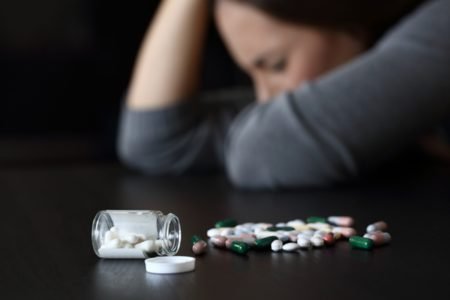 Imagem colorida: remédios sobre a mesa e mulher no fundo com a mão na cabeça - Metrópoles