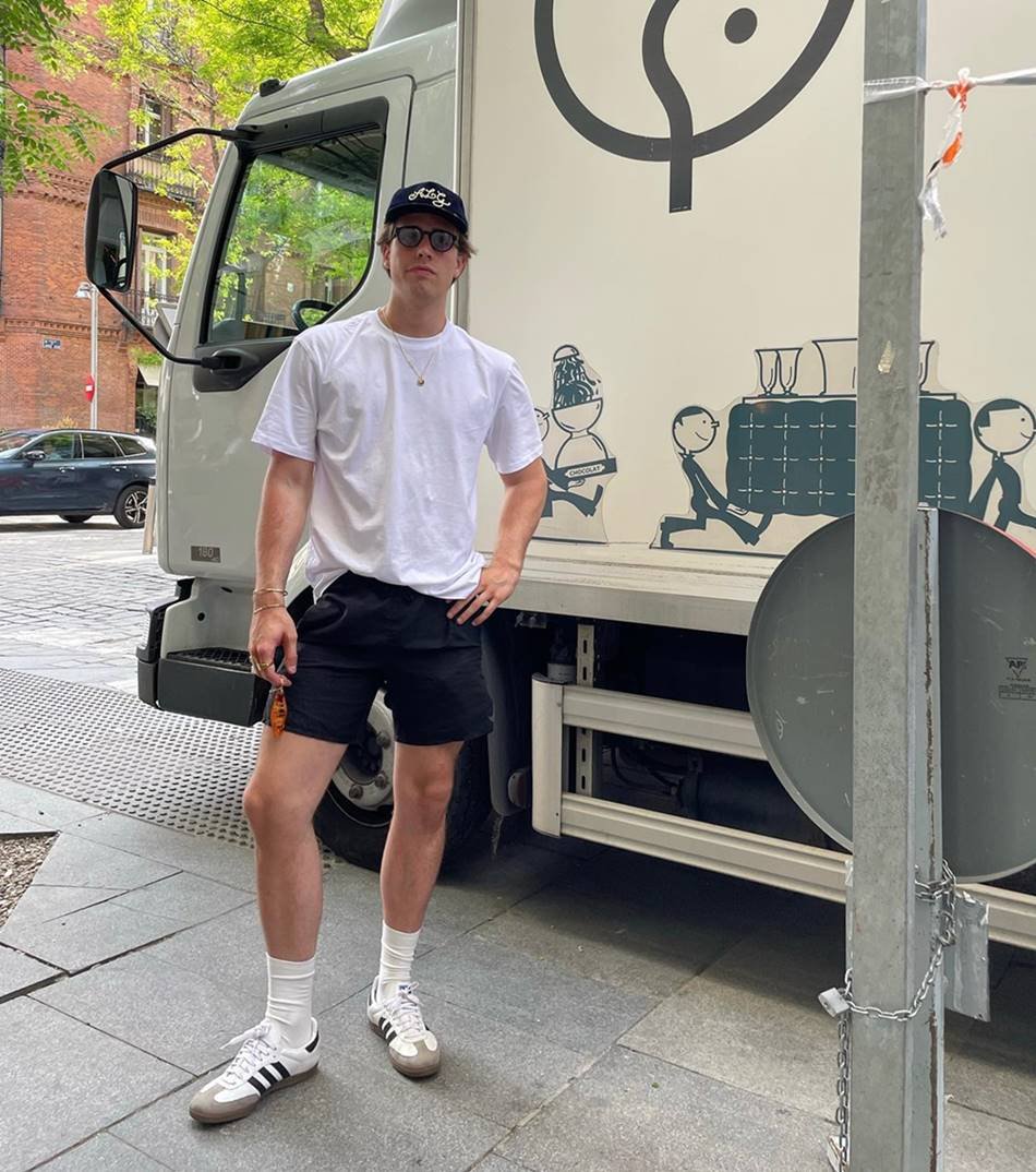 Homem jovem e branco, com cabelo loiro liso, posa para foto ao lado de um caminhão, em Nova York. Usa uma camiseta branca, um short preto, tênis Samba da marca Adidas e boné preto. - Metrópoles