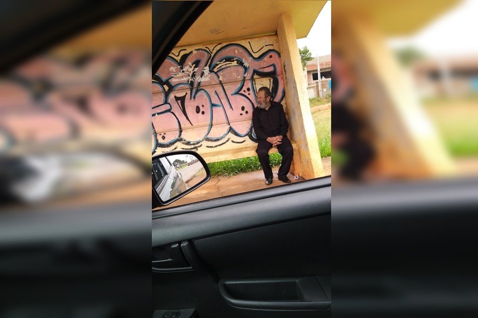 José Tavares sentado em uma parada de ônibus com roupa social preta