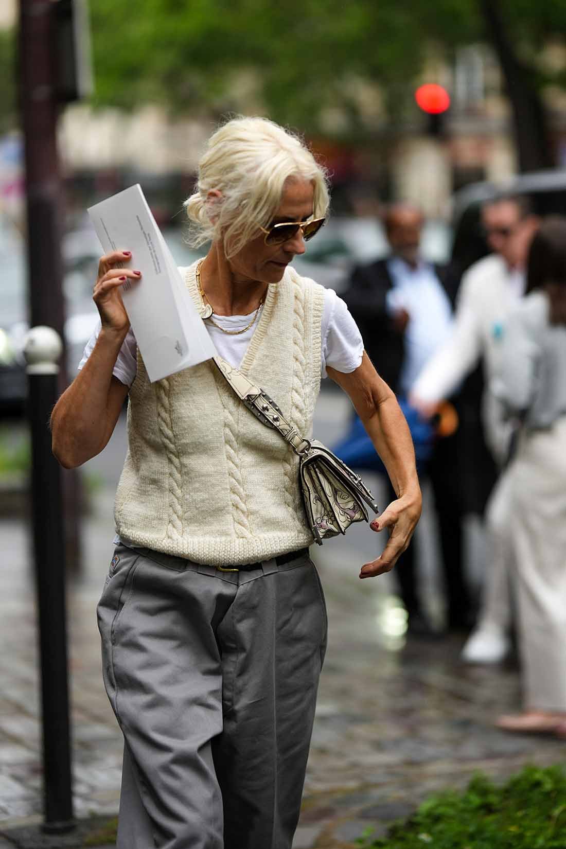 Mulher branca e idosa, de cabelos lisos e brancos, anda pela rua durante a Semana de Moda de Paris. Ela usa uma camiseta branca, um colete de tricô bege e calça de alfaiataria cinza, além de óculos escuros. - Metrópoles