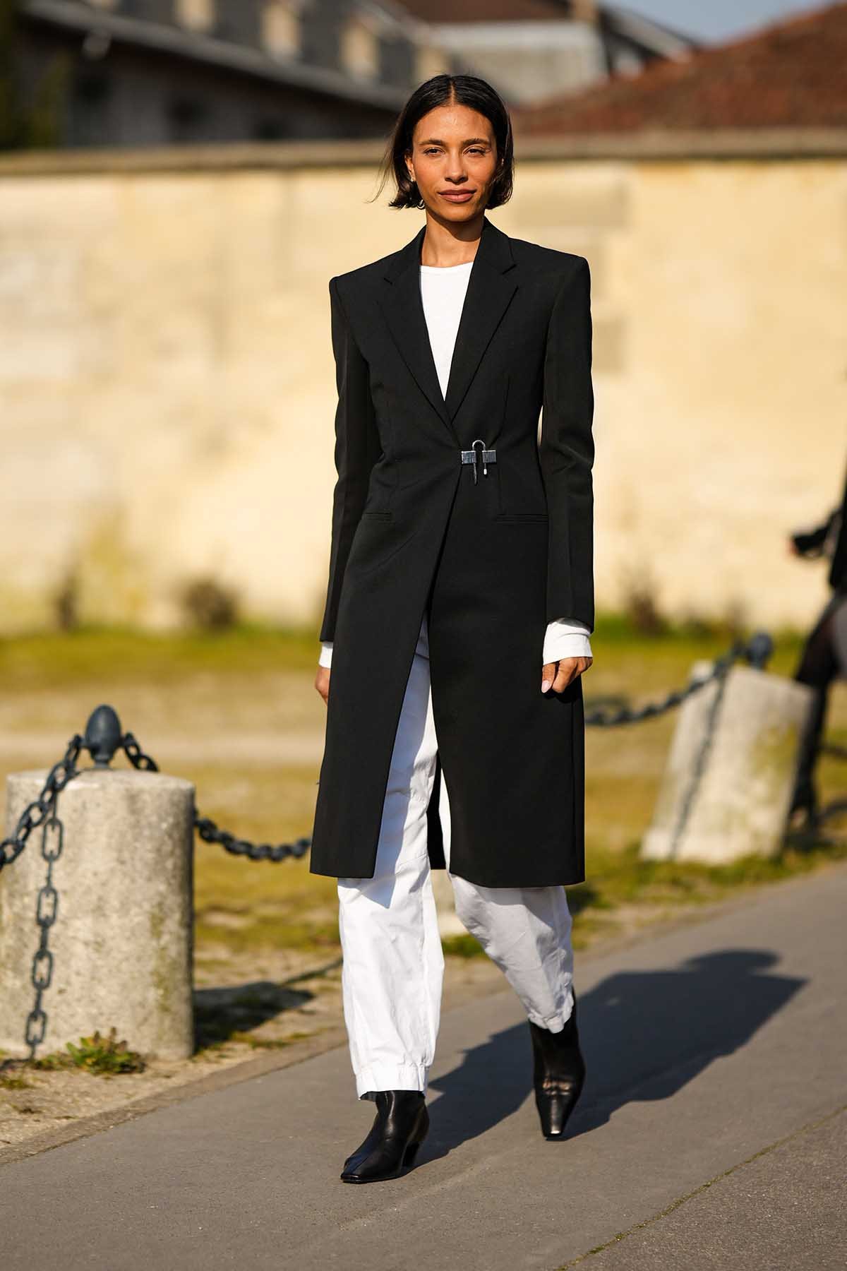 Mulher morena e jovem, de cabelo preto liso, andando pela rua durante a Semana de Moda de Paris. Ela usa uma camiseta e uma calça, ambas brancas; um casaco longo preto de alfaiataria e uma bota de couro preta. - Metrópoles