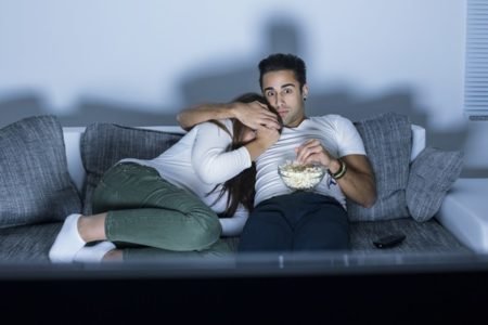 Imagem colorida de casal sentado no sofá assistindo a filme de terror - Metrópoles