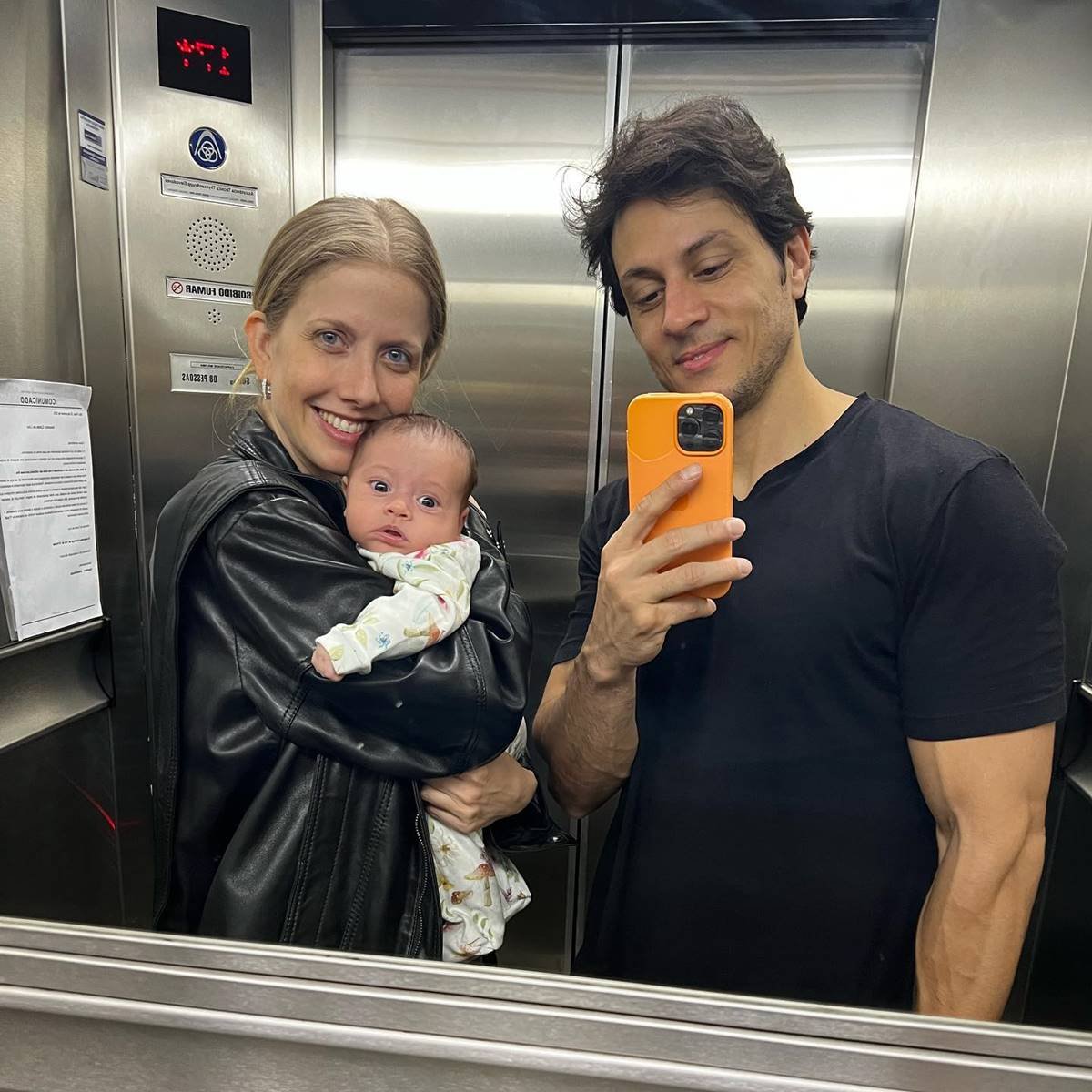 A apresentadora Gabriela Prioli, uma mulher branca e loira, posa para foto no espelho de um elevador segurando sua filha Ava. Ela está ao lado do marido, o DJ Thiago Mansur. - Metrópoles