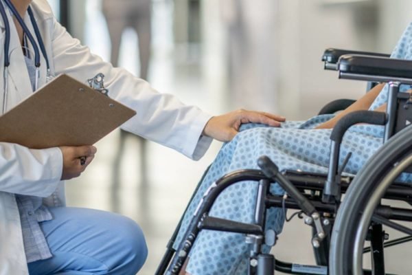 Médica de joelhos em frente a paciente em cadeira de rodas