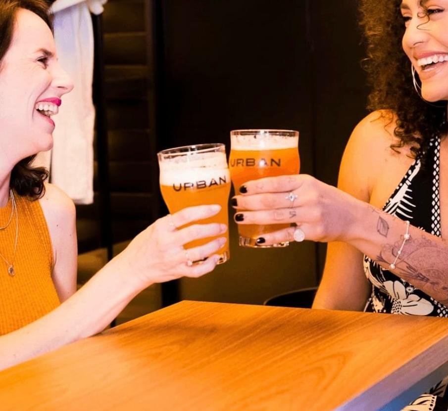 Foto de duas mulheres brindando um copo de cerveja enquanto se olham e sorriem 