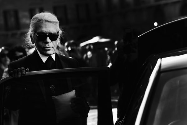 O estilista Karl Lagerfeld, um homem branco e idoso, de cabelos lisos brancos, entrando em um carro. Ele usa uma camisa de botão branca e colete e calça pretos. A foto é preta e branca. - Metrópoles