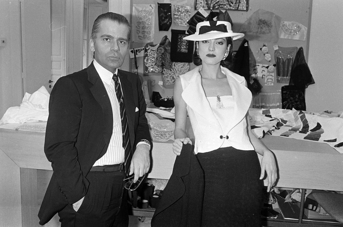 O estilista Karl Lagerfeld ao lado de uma modelo em desfile da grife Chanel, em 1983. Ele usa uma camisa, um blazer e uma gravata e ela um vestido preto e branco e chapéu. Ambos são brancos e jovens. - Metrópoles