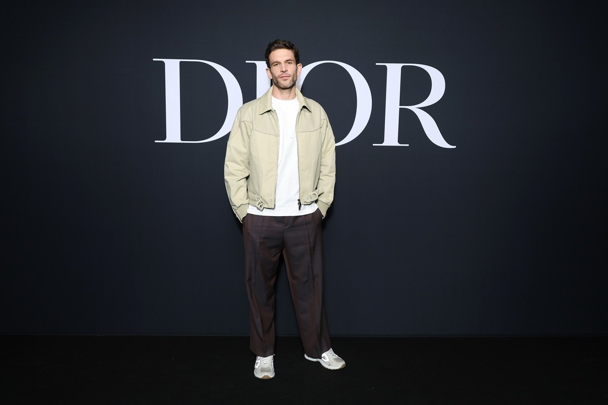 O ator Arnaud Valois, um homem branco e jovem, de cabelo castanho, no desfile da Dior. Ele usa uma camiseta branca, um casaco bege e uma calça preta. - Metrópoles