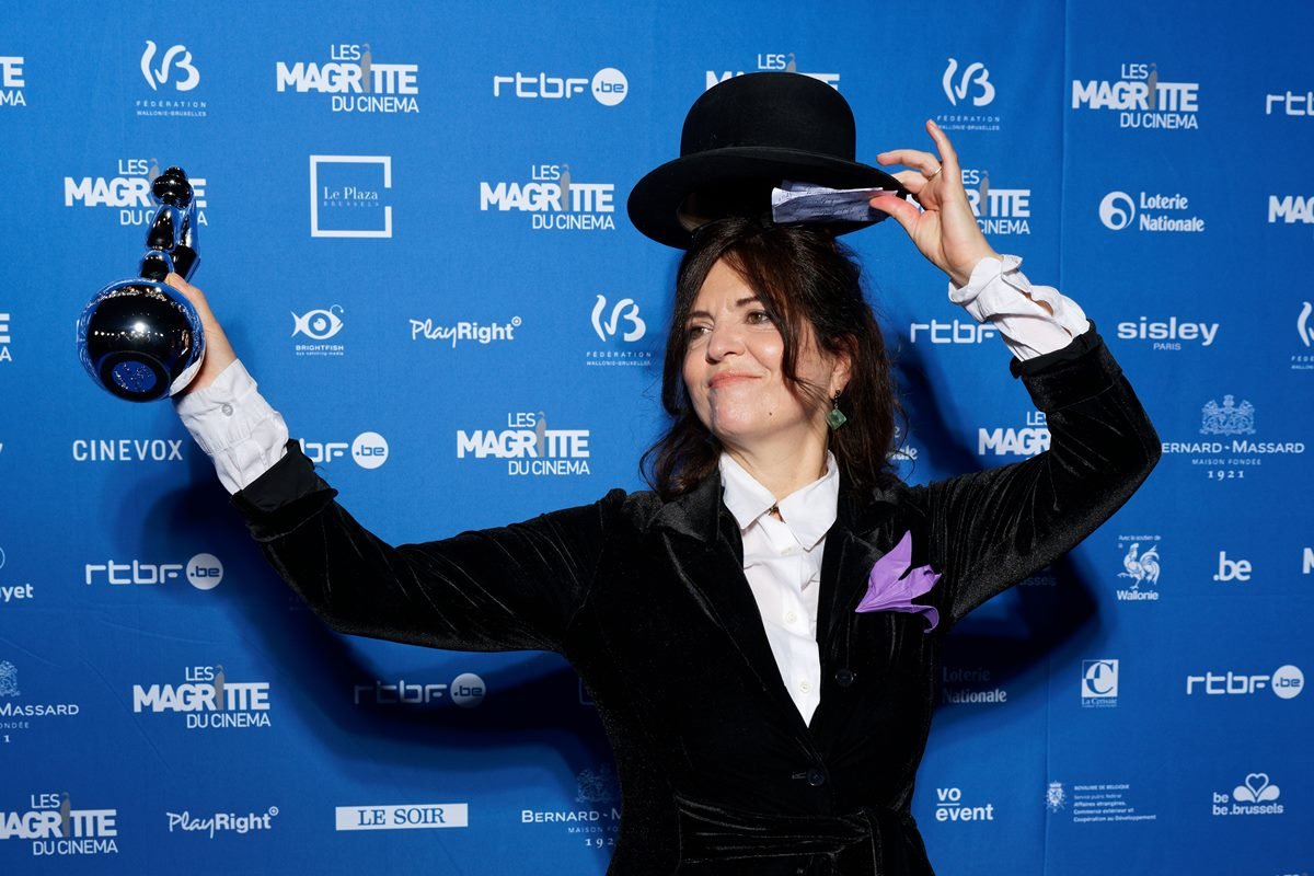 A atriz Agnès Jaoui, uma mulher branca e de meia idade, com cabelo preto ondulado, ela usa uma camisa branca e um casaco preto de camurça, além de um chapéu preto de coco. - Metrópoles