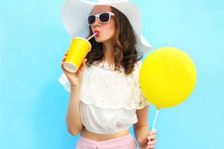 mulher segurando um balão amarelo e tomando algo no copo amarelo de óculos e chapéu
