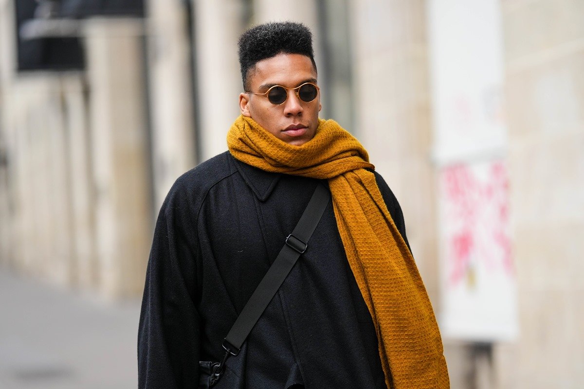 Homem negro e jovem, de cabelo cacheado curto, anda pela rua durante a Semana de Moda de Paris. Ele usa um casaco preto e um cachecol mostarda, além de óculos escuros e uma bolsa transversal. - Metrópoles