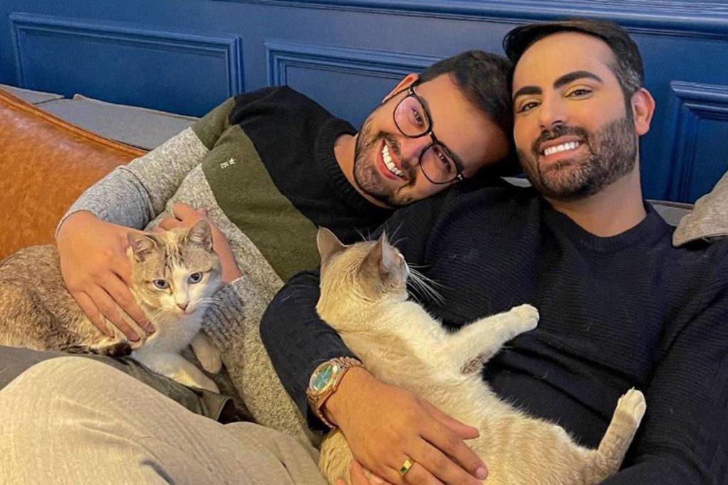 dois homens sentados sorrindo em sofá com gatos no colo - metrópoles
