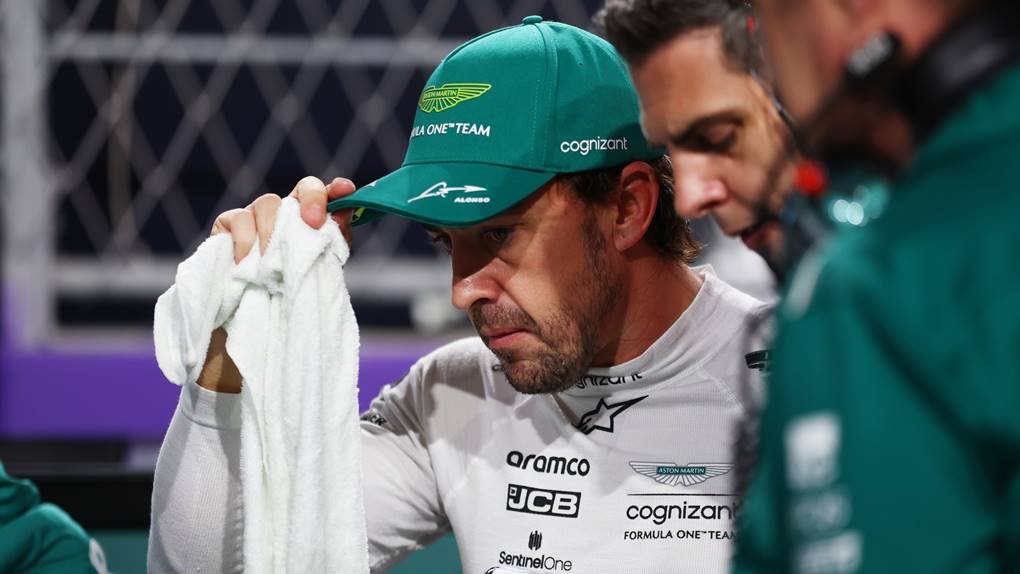 Foto do piloto Fernando Alonso com macacão branco, com semblante triste e colocando a mãe em seu boné que está na cabeça