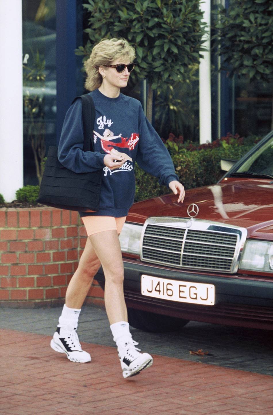 Foto da princesa Diana andando pelas ruas de Londres em 1995. Ela usa um casaco de moletom azul, short de ciclista rosa, tênis esportivo branco e um óculos de sol. - Metrópoles